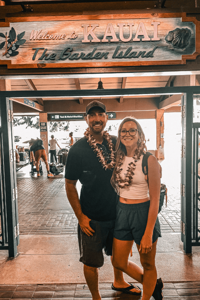 Kate and her companion stand under a “Welcome to Kauai – The Garden Island” sign at the airport, smiling with fresh leis around their necks as they begin their Hawaiian adventure.