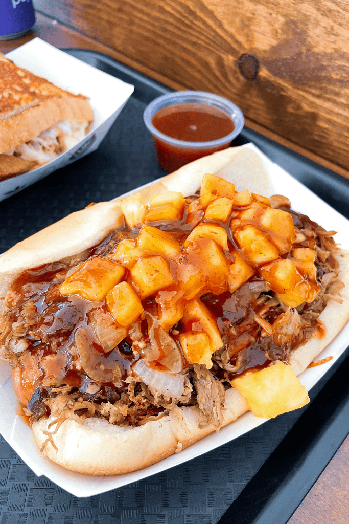 Close-up of a juicy pulled pork sandwich topped with grilled onions, pineapple chunks, and a generous drizzle of barbecue sauce from Porky’s Kauai.