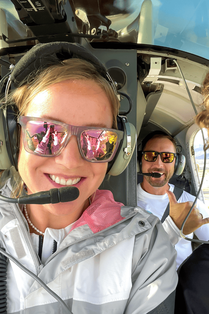 Kate and her husband smile wide in a helicopter selfie, both wearing aviation headsets and sunglasses, excited for their aerial adventure over Kauai.