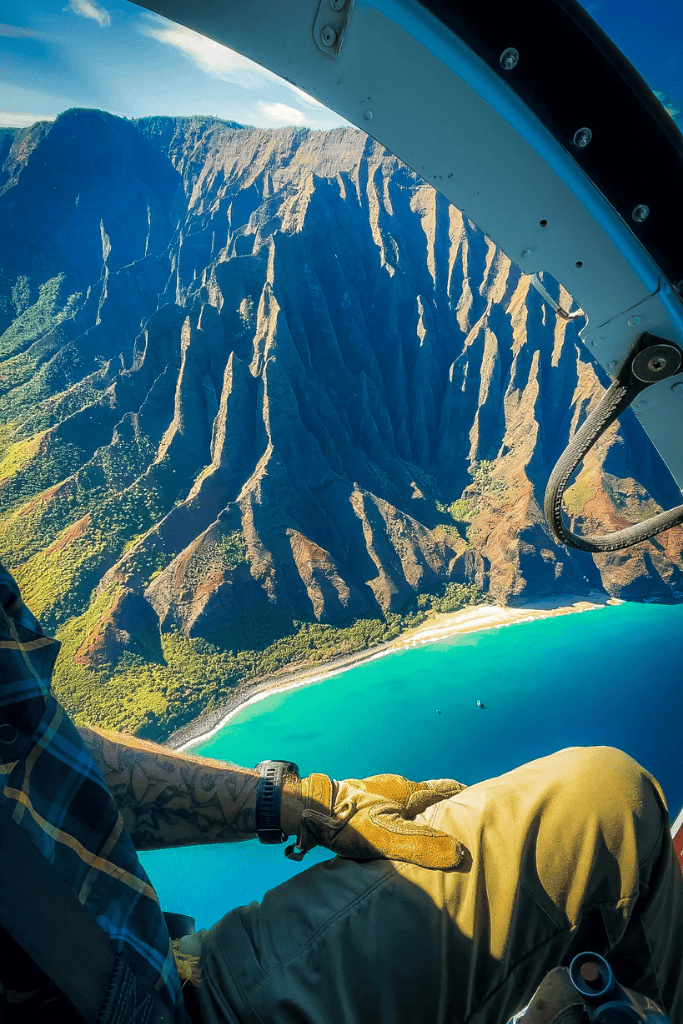 Stunning aerial view of the Nā Pali Coast from the open door of a helicopter, revealing dramatic cliffs and turquoise water below.
