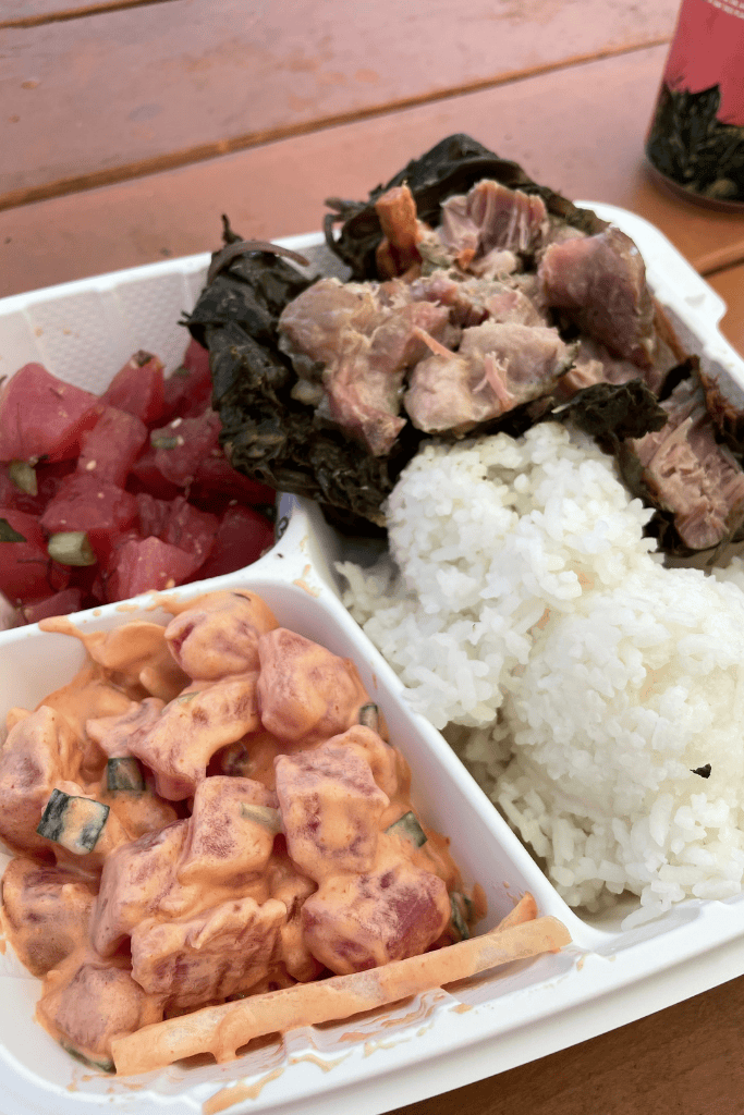A Hawaiian plate lunch featuring lau lau pork, white rice, spicy ahi poke, and shoyu poke served in a takeout container on a picnic table.