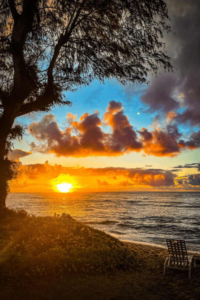 A lone beach chair faces the Pacific Ocean beneath golden clouds as the sun rises dramatically from the horizon in Kauai, framed by trees and glowing foliage.