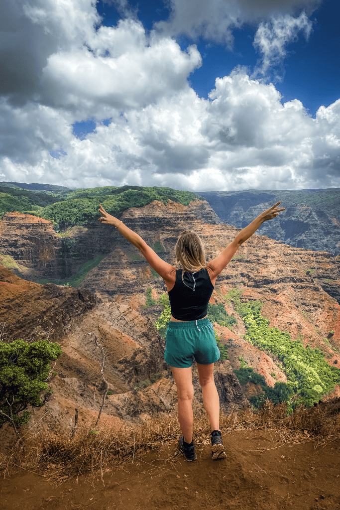 Kate celebrates the breathtaking view at Waimea Canyon, with arms raised over a vast landscape of layered red rock and lush green valleys.