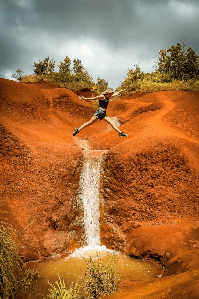 Mid-leap over the iconic Waimea Canyon red dirt waterfall, Kate’s athletic jump adds energy and excitement to the vividly colored terrain under a dramatic sky.
