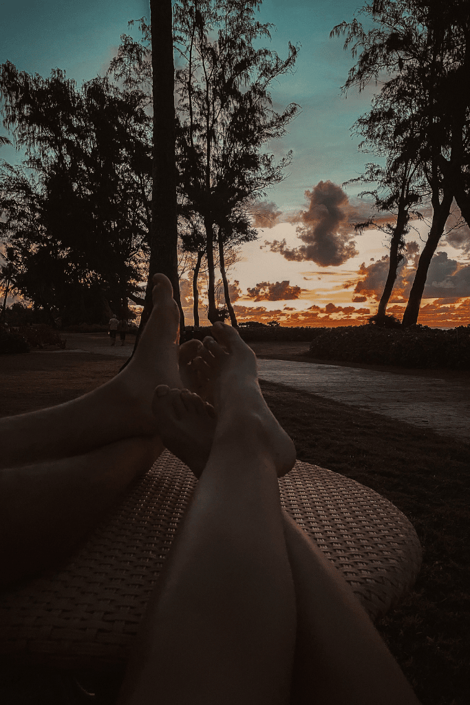 With their feet up on a wicker table, Kate and her husband unwind beneath silhouetted trees, watching the final glow of sunset paint the Kauai sky in hues of orange and blue.