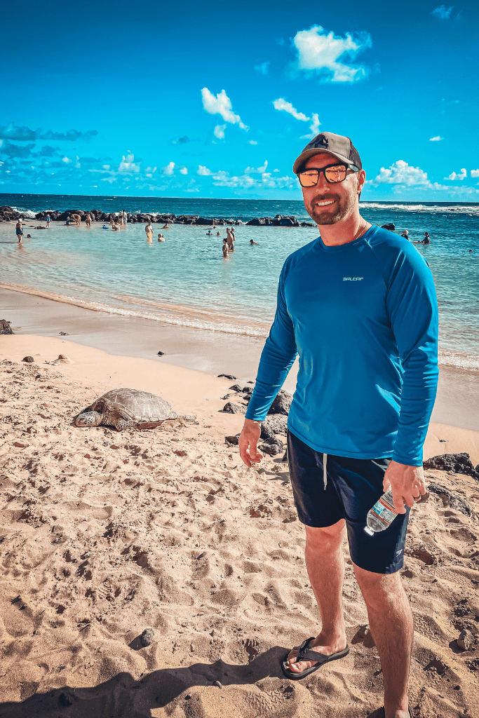 Kate's husband smiles on a sunny Kauai beach with a sea turtle in the sand nearby, as swimmers enjoy the turquoise waters in the background.
