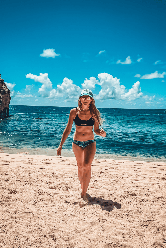 Kate, in a black bikini and teal hat, walks barefoot on the sandy shore of Larsen’s Beach, with a dramatic blue sky and ocean stretching out behind her.