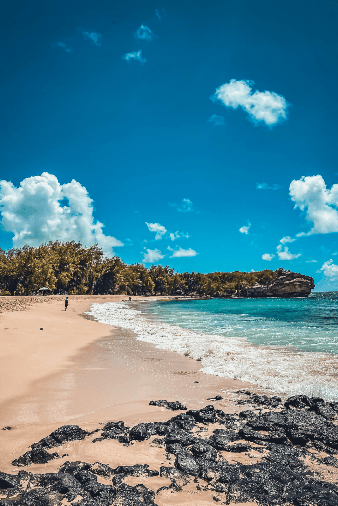 A wide sandy stretch of Shipwreck Beach in Kauai curves along turquoise waters under a bright blue sky, with a rocky cliff jutting out at the far end and only a few people scattered along the serene shoreline.