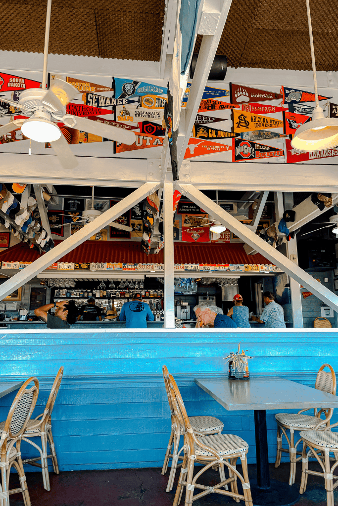 Interior of Brennecke’s Beach Broiler on Kauai, featuring bright blue walls, a bar lined with license plates and collegiate pennants, and hanging hats from the ceiling.