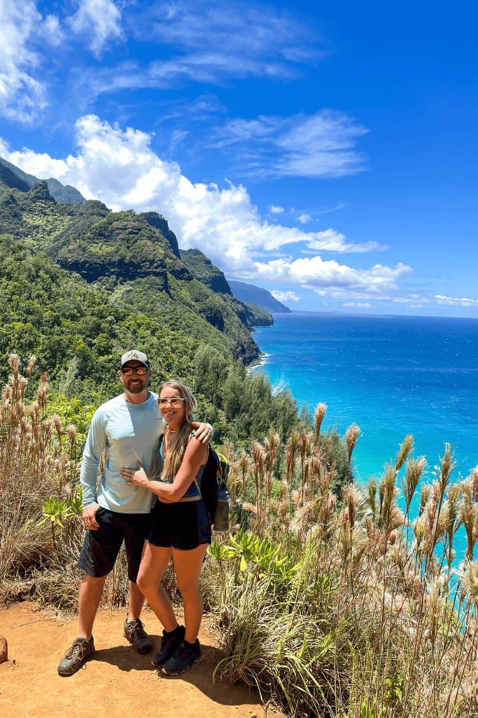 Kate and her husband pose on the Kalalau Trail, surrounded by lush jungle and overlooking the turquoise waters and rugged cliffs of Kauai’s Nā Pali Coast.