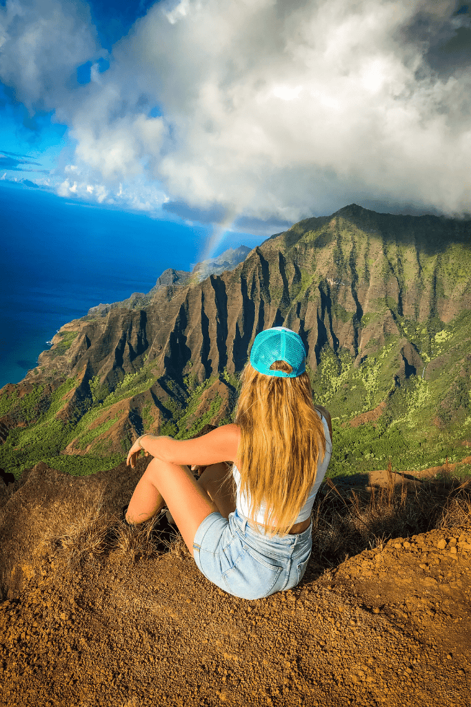Kate takes a peaceful seat at the cliff’s edge, soaking in the panoramic view of the Nā Pali Coastline, where sunlight and clouds dance over the dramatic ridges and a rainbow arcs in the distance.