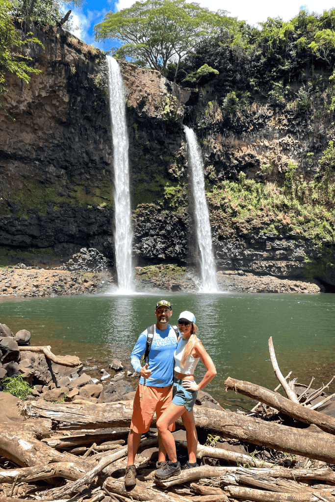 Kate and her companion pose in front of the iconic twin streams of Wailua Falls, surrounded by driftwood and lush cliffside greenery.