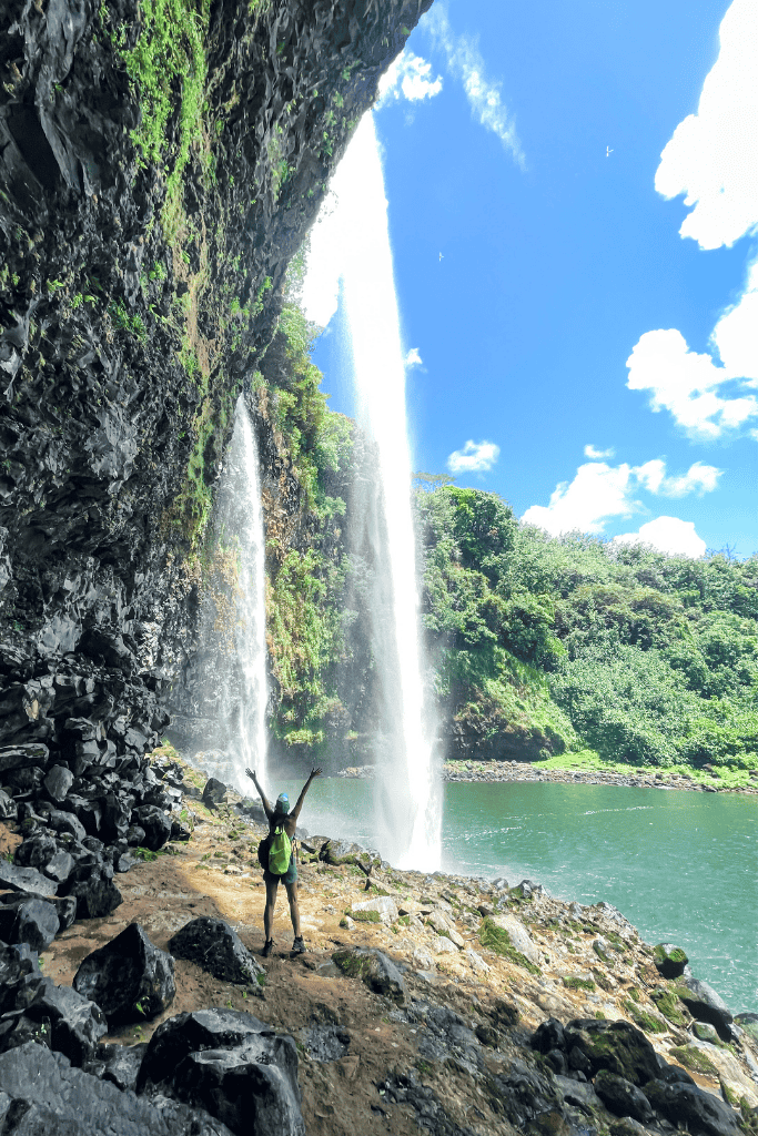 A triumphant Kate raises her arms beneath the misty spray of Wailua Falls, viewed dramatically from behind the waterfall.