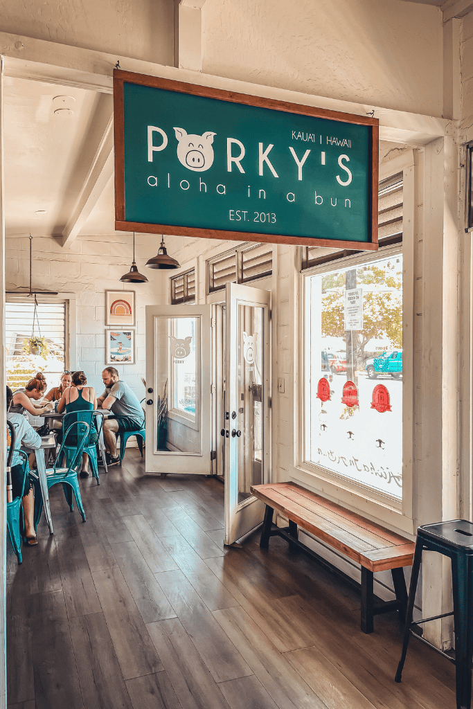 Interior of Porky’s Kauai, a laid-back eatery with a teal sign that reads “Aloha in a Bun,” as diners enjoy their meals at rustic tables.