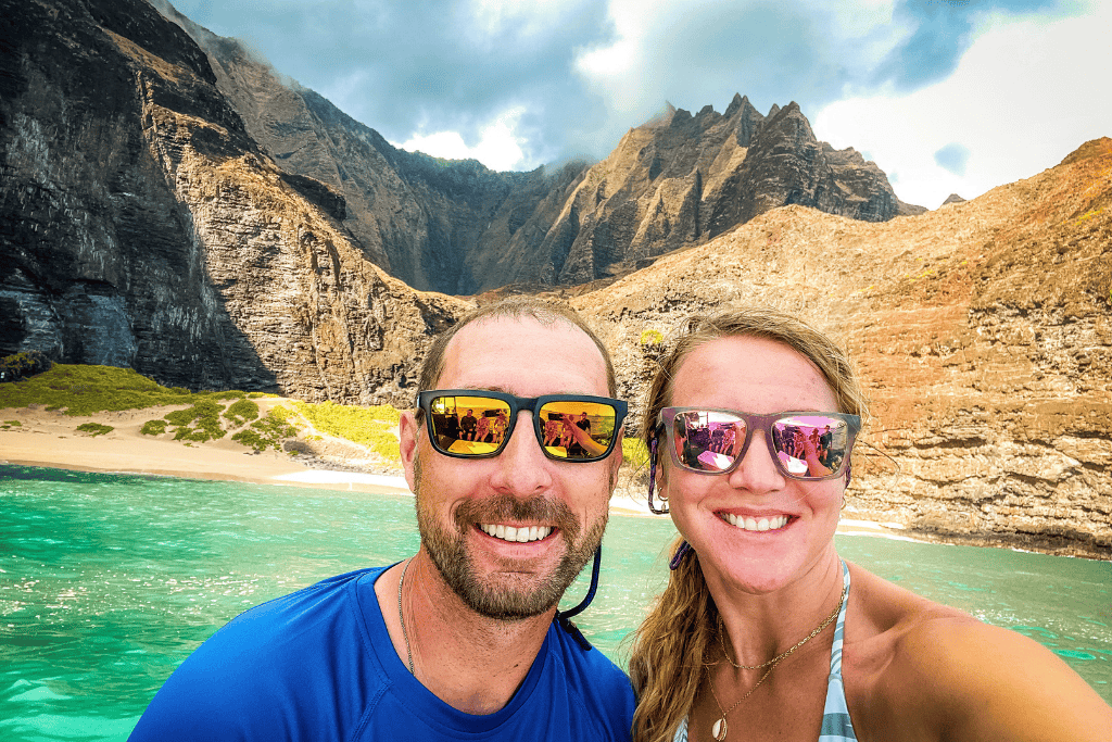 Kate and her husband take a vibrant boat selfie with the jagged cliffs of the Nā Pali Coast rising dramatically behind them, their sunglasses reflecting the stunning turquoise waters.
