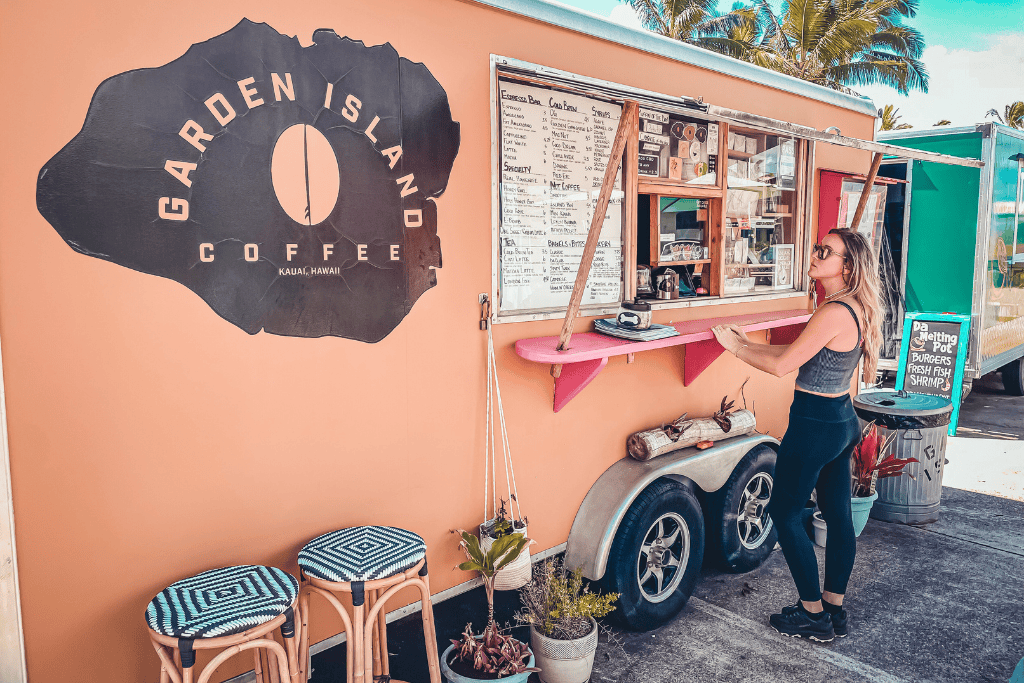 Kate stands at the window of the vibrant Garden Island Coffee truck in Kauai, reading the menu under swaying palms with island breeze in the air.