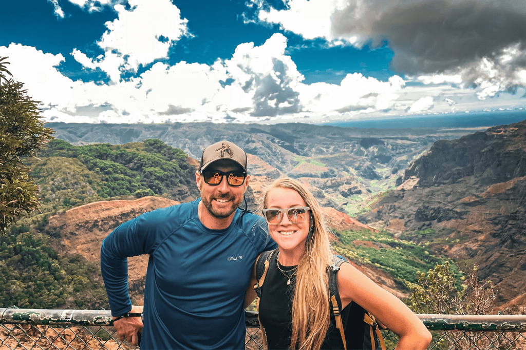 Smiling against the sweeping backdrop of Waimea Canyon, Kate and her husband pose at a scenic lookout, the vibrant reds and greens of the landscape stretching into the distance beneath a dramatic sky.
