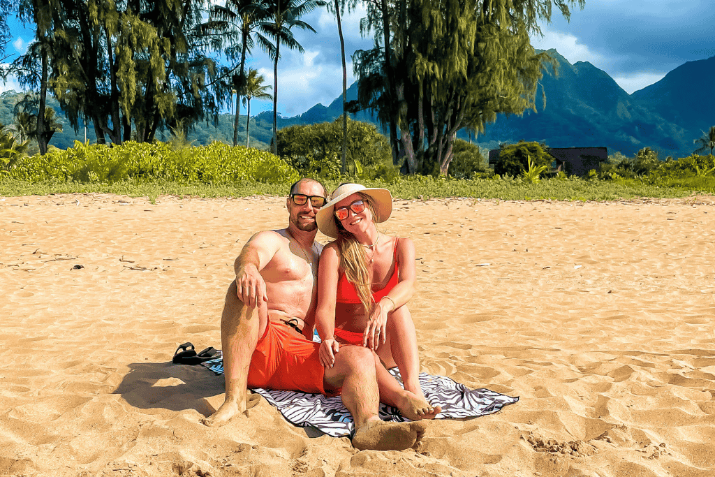 Kate and her husband relax together on the golden sands of Hanalei Bay, basking in the sun with the lush green mountains of Kauai rising majestically in the background.