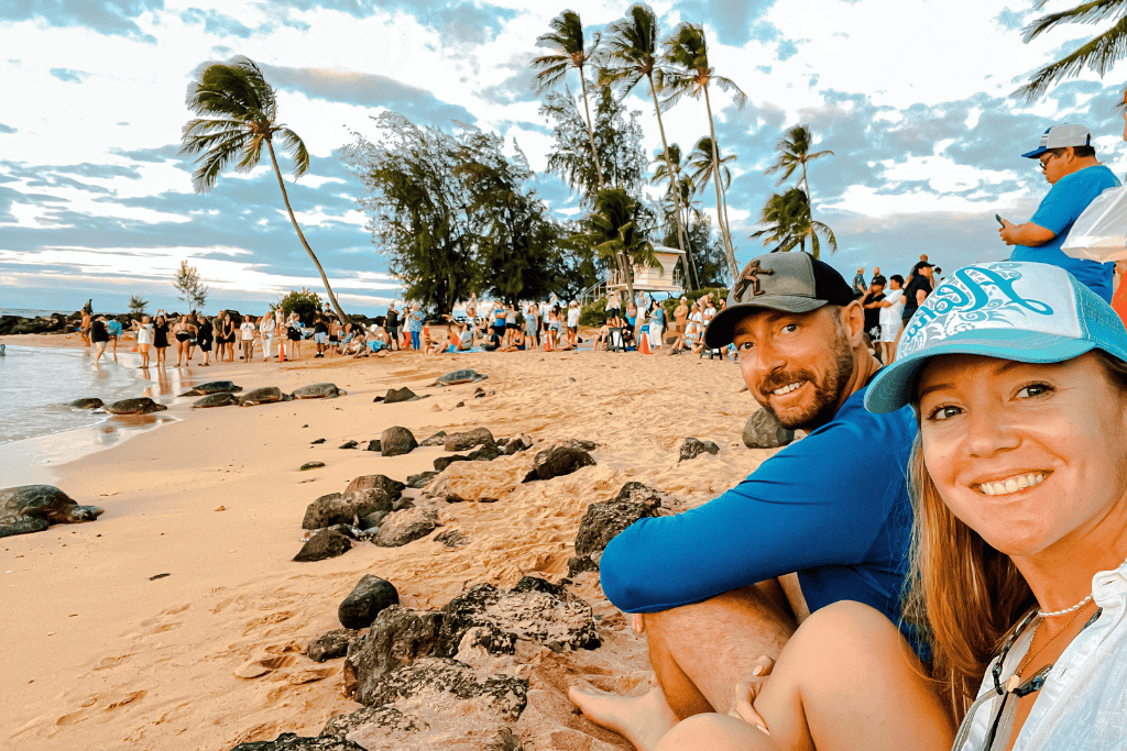 Kate and her husband smile for a sunset selfie on Poipu Beach as a crowd gathers behind them to watch Hawaiian sea turtles haul out onto the shore for the evening.