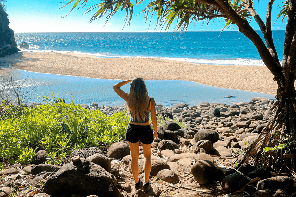 Kate stands on a rocky trail framed by tropical foliage, gazing out at the golden sands and bright blue waters of Hanakapiai Beach on Kauai’s North Shore.