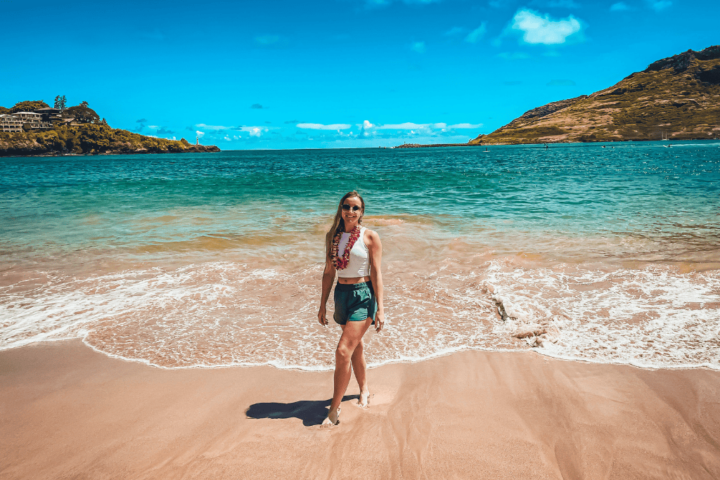 Kate walks barefoot along the shoreline of Kalapaki Beach, wearing sunglasses and a fresh orchid lei as gentle waves roll in under a sunny Kauai sky.