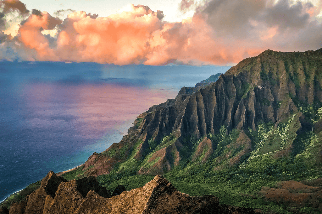 Stunning view of the Nā Pali Coast at sunset, with jagged green cliffs plunging into the Pacific Ocean and golden clouds casting a warm glow across the water.