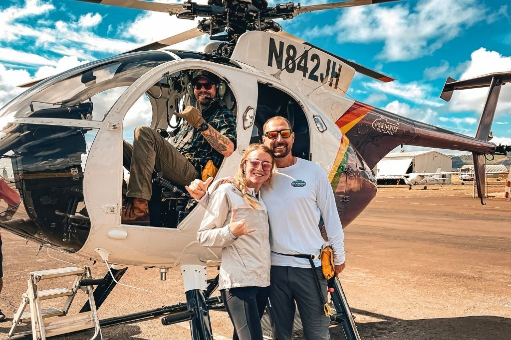 Kate and her husband posing in front of a helicopter with the pilot seated inside, giving a thumbs-up. The helicopter features a colorful design and sits on a sunny tarmac with a blue sky and hangars in the background, capturing the excitement of an adventurous helicopter tour in Hawaii.