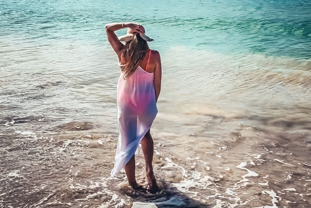 This image shows Kate standing in shallow ocean water, with their back turned towards the camera, wearing a sheer white dress that is illuminated by sunlight. The setting captures a peaceful beach moment, with clear turquoise water in the background.