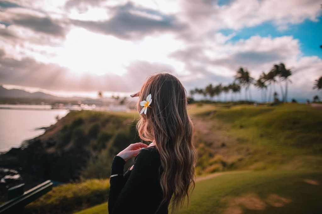 This image captures a person with long wavy hair adorned with a white plumeria flower, standing on a grassy landscape overlooking a coastal view. The scene is bathed in soft, golden light with palm trees in the background and dramatic clouds scattering sunlight across the sky