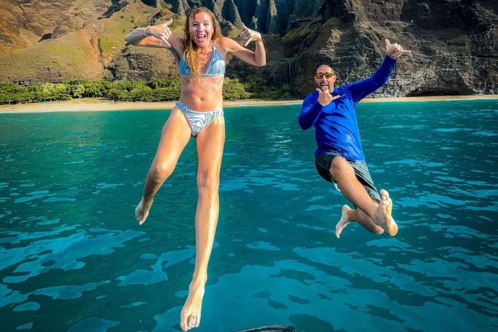 This image captures Kate and her husband mid-air as they jump into clear turquoise water, with dramatic cliffs and lush greenery of Kauai in the background. Kate on the left is wearing a patterned bikini, and her husband on the right is in a blue rash guard, both radiating excitement and energy.