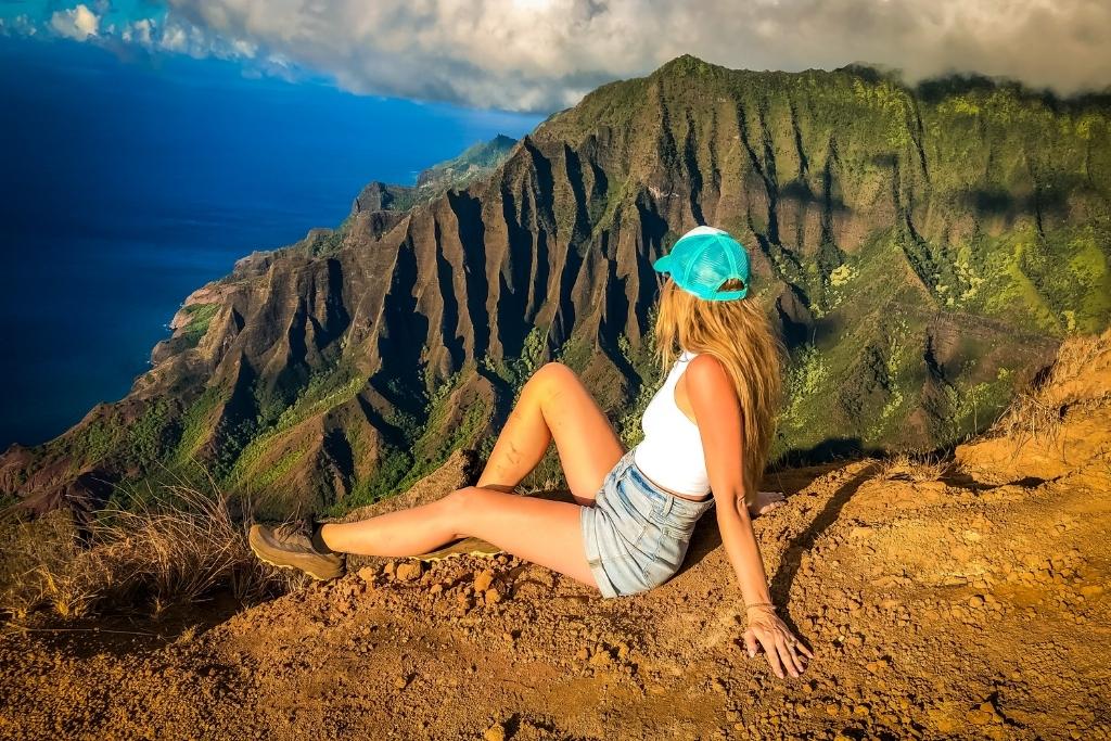 This image Kate sitting on the edge of a cliff overlooking the breathtaking Na Pali coastline in Kauai, with its dramatic green ridges and deep valleys meeting the ocean. The person is wearing a blue cap, white tank top, denim shorts, and hiking shoes, basking in the golden sunlight.