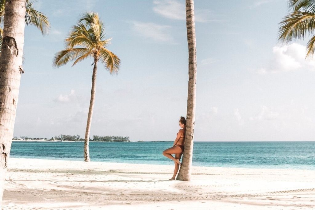 This image features a person leaning against a palm tree on a pristine white sandy beach, wearing a swimsuit and enjoying the serene tropical setting. The turquoise ocean stretches out in the background, bordered by a distant island and clear skies dotted with soft clouds.