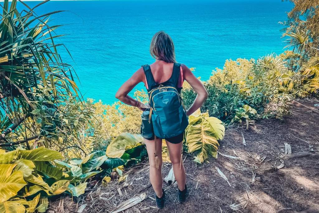 This image shows Kate standing on a lush trail overlooking a vibrant turquoise ocean, wearing a backpack and outdoor gear. Surrounded by tropical foliage, they appear to be enjoying the stunning coastal view, capturing the essence of a hiking adventure.