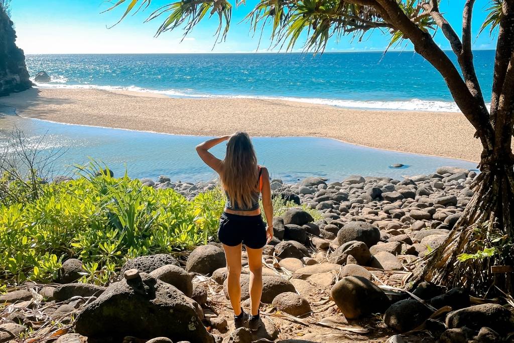 This image shows Kate standing on a rocky trail framed by tropical greenery, overlooking a serene beach with golden sand and gentle waves meeting the shore. The scene is bathed in bright sunlight, with the ocean stretching out under a clear blue sky.