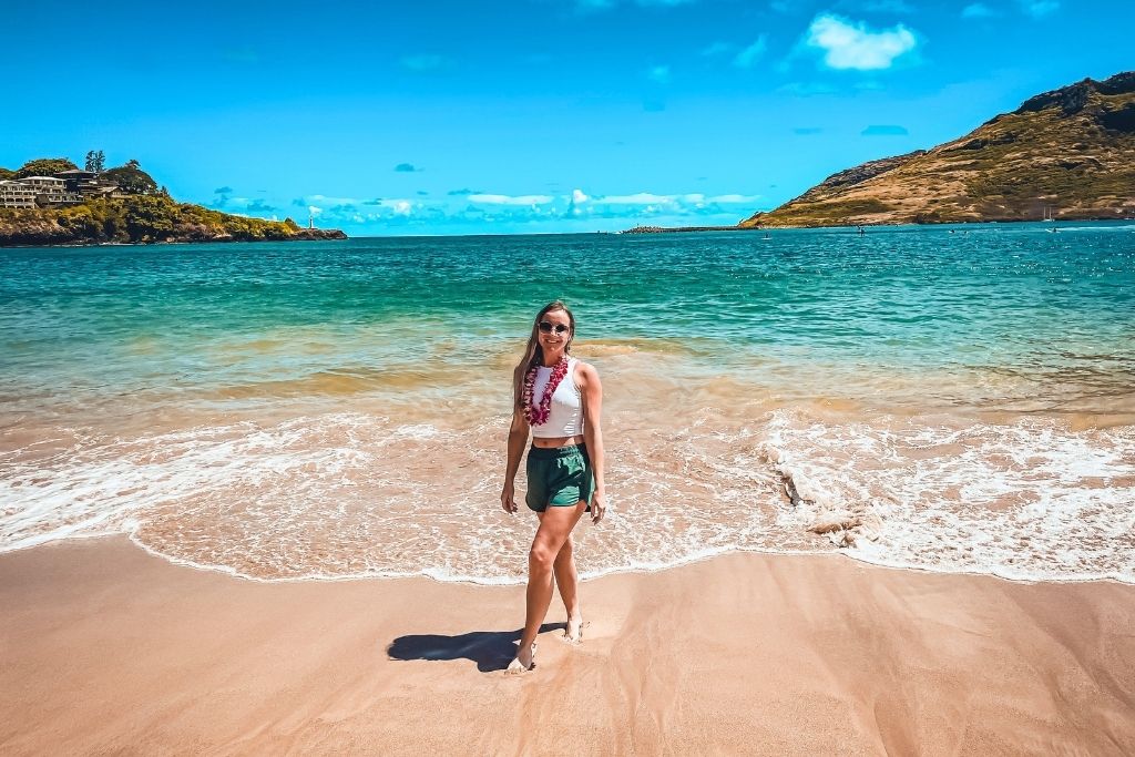 Kate wearing a lei and summer outfit standing on a sandy beach with clear turquoise waters and green hills in the background. Perfect depiction of Hawaii beach vibes, ideal for a Hawaii packing list that includes lightweight clothing, beach essentials, and accessories for a beach vacation.