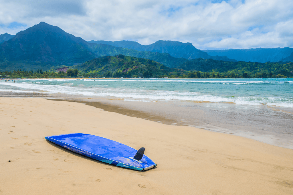 A bright blue surfboard lies fin-up on the golden sand of Hanalei Bay, with gentle waves and lush Kauai mountains in the background.