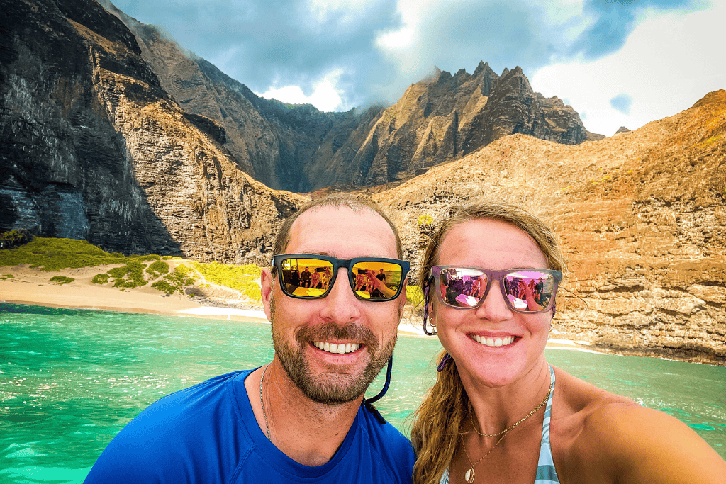 Kate and her companion smile for a selfie in front of the dramatic cliffs and turquoise waters of Kauai’s Nā Pali Coast, both wearing sunglasses that reflect the stunning scenery.