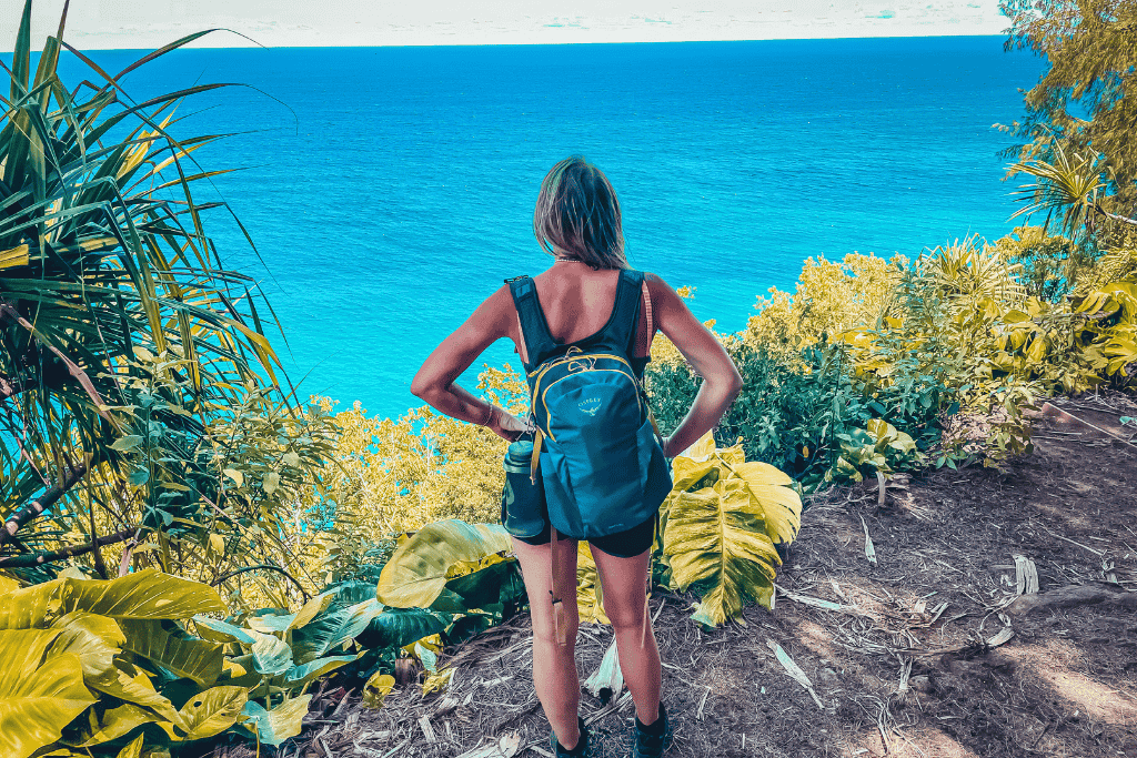 Kate stands at a lush cliffside viewpoint with hands on hips, wearing a backpack and gazing out at the endless blue expanse of the Pacific Ocean in Kauai.