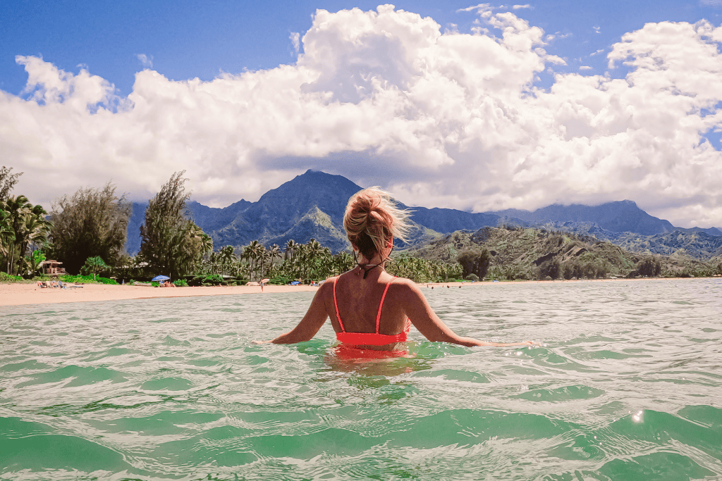 Kate floats in waist-deep emerald water facing the shore, with Hanalei Bay’s lush mountains and dramatic clouds rising in the distance.