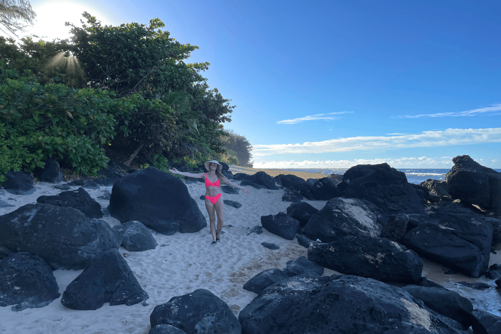 Kate stands on a secluded sandy beach in a bright pink bikini and sunhat, surrounded by large black lava rocks and lush tropical greenery under a clear blue sky in Kauai.