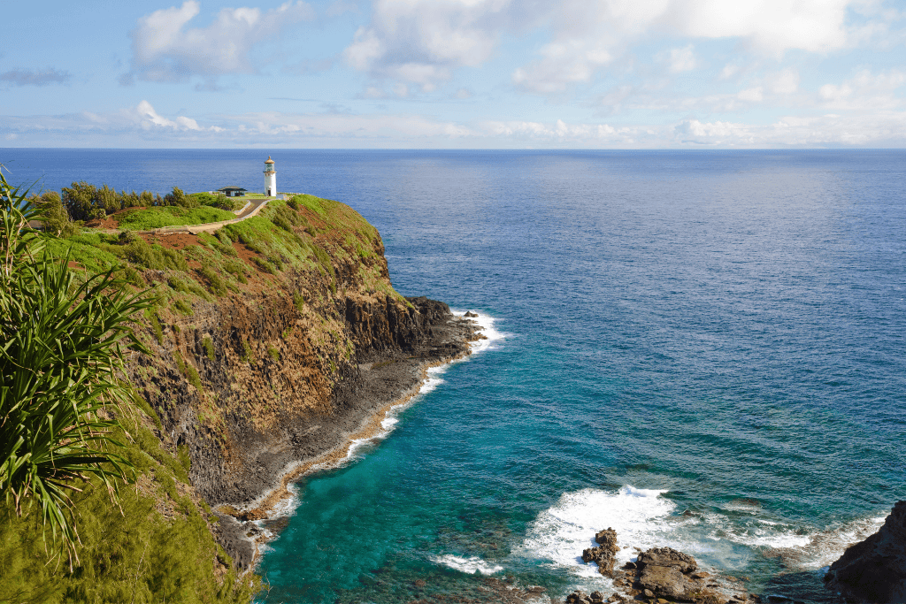 A white lighthouse stands on the edge of a steep, grassy cliff overlooking the deep blue Pacific Ocean at Kīlauea Point on Kauai’s northern coast.