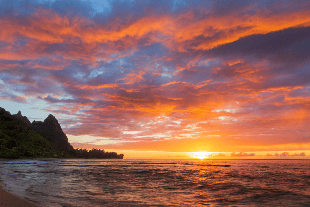 A vibrant Kauai sunset paints the sky in fiery shades of orange, pink, and purple over the ocean, with a dark silhouette of rugged cliffs lining the shore.