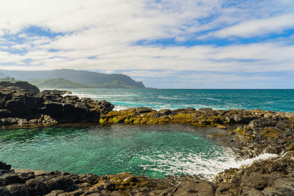 Natural tidepool surrounded by black lava rock with turquoise water and ocean waves in the background, taken on the north shore of Kauai.