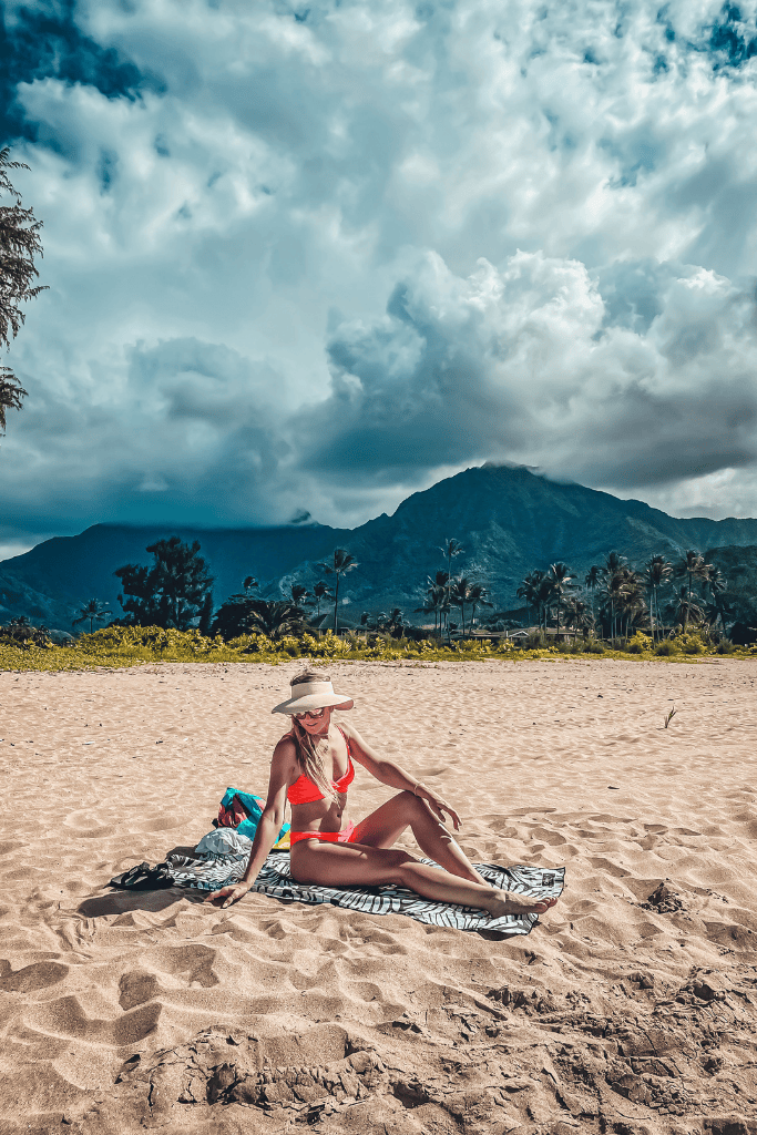 Kate lounges on a beach towel in a red bikini and sunhat, with dramatic clouds and the mountains of Kauai rising behind her.