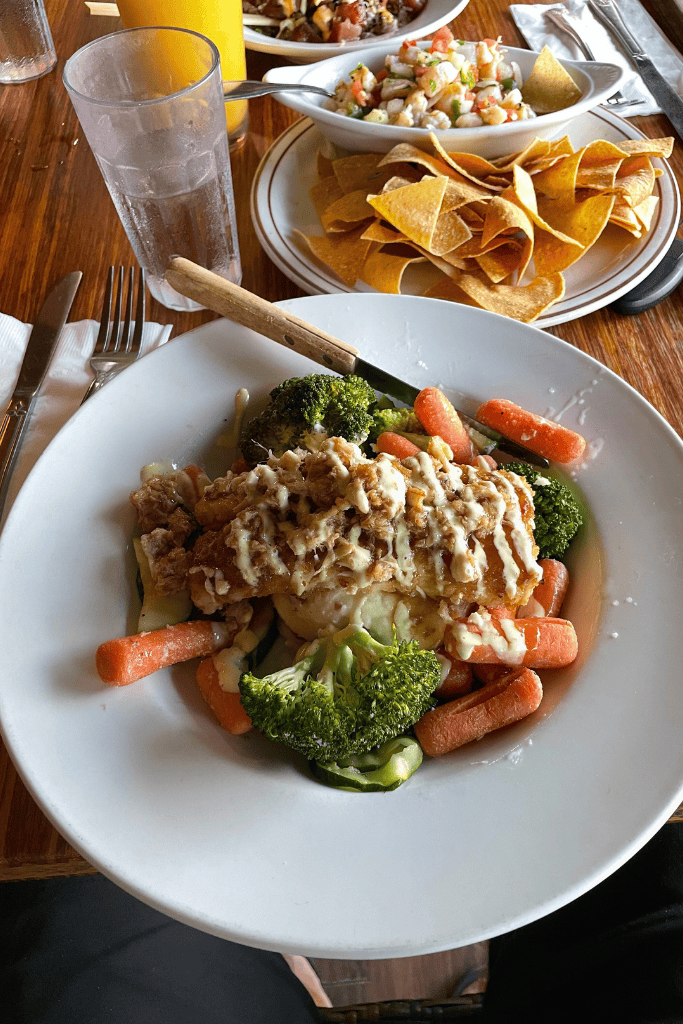 A plate of crispy fish topped with creamy sauce and chopped nuts, served over sautéed vegetables including broccoli and carrots, with chips, salsa, and shrimp ceviche in the background.
