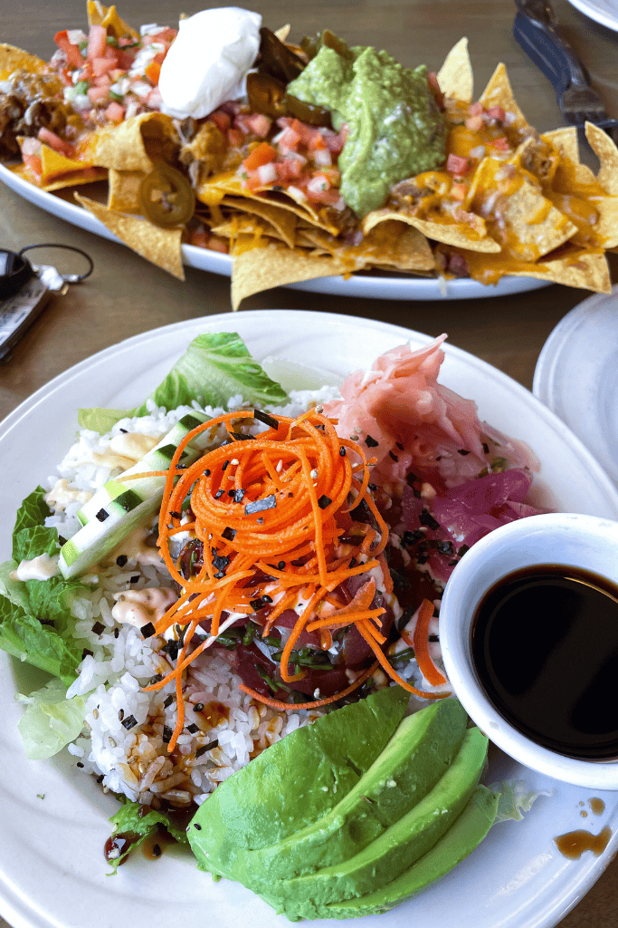 A fresh poke bowl with rice, ahi tuna, shredded carrots, avocado, cucumber, and pickled ginger, served alongside a soy dipping sauce and a plate of loaded nachos.