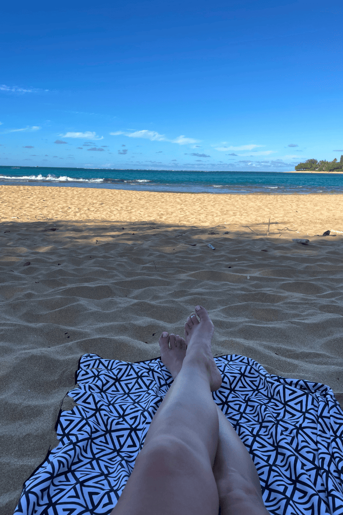 Kate’s legs are stretched out on a patterned beach towel facing the ocean, with soft golden sand and gentle waves under a clear Kauai sky.