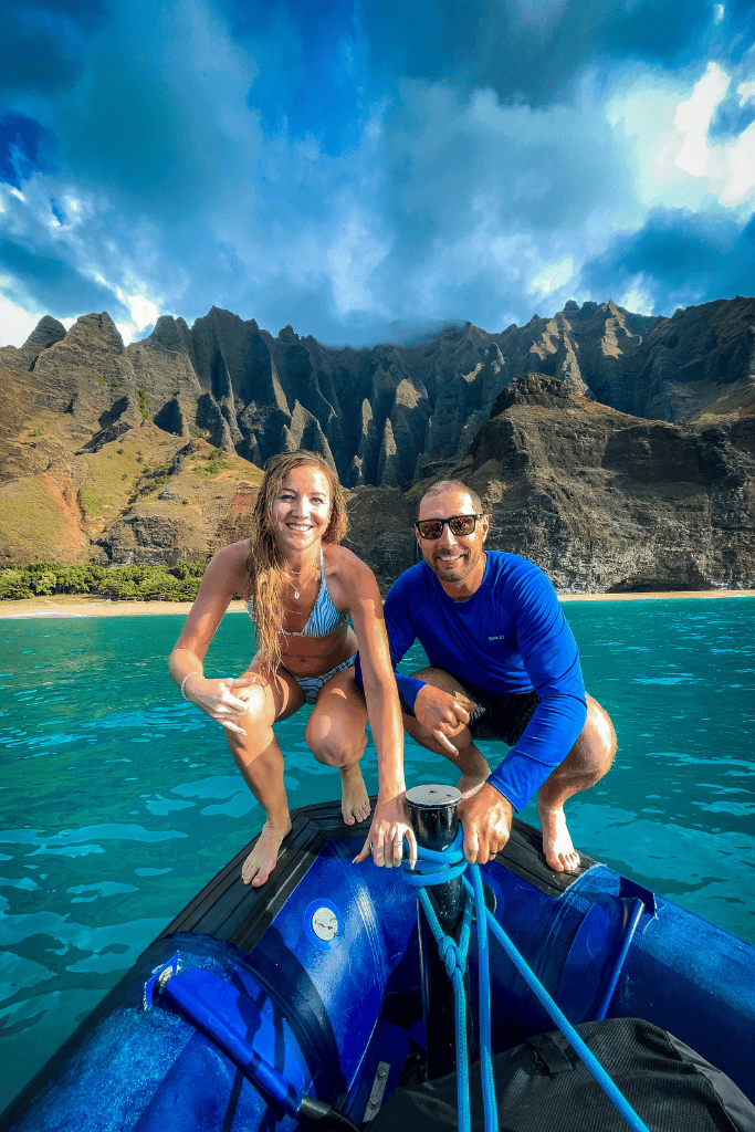 Kate and a man smile on the edge of a small boat in front of the dramatic cliffs of Kauai’s Nā Pali Coast, with turquoise water beneath them.
