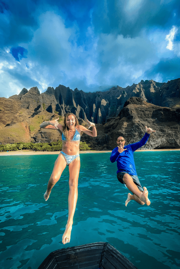 Mid-jump, Kate and her companion leap from their boat into the ocean, framed by the towering peaks of the Nā Pali Coast under a vivid sky.