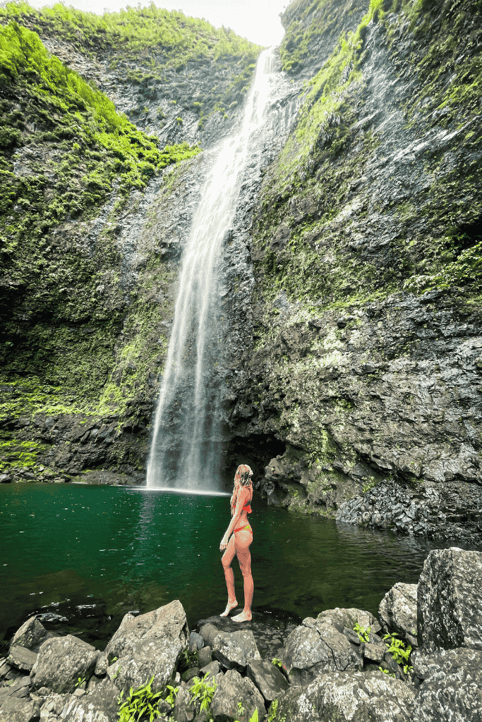 Kate stands barefoot in a red bikini at the edge of a jade-green pool, admiring a towering waterfall framed by steep, mossy cliffs.